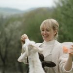A smiling woman feeds a goat outdoors, capturing a peaceful countryside moment.